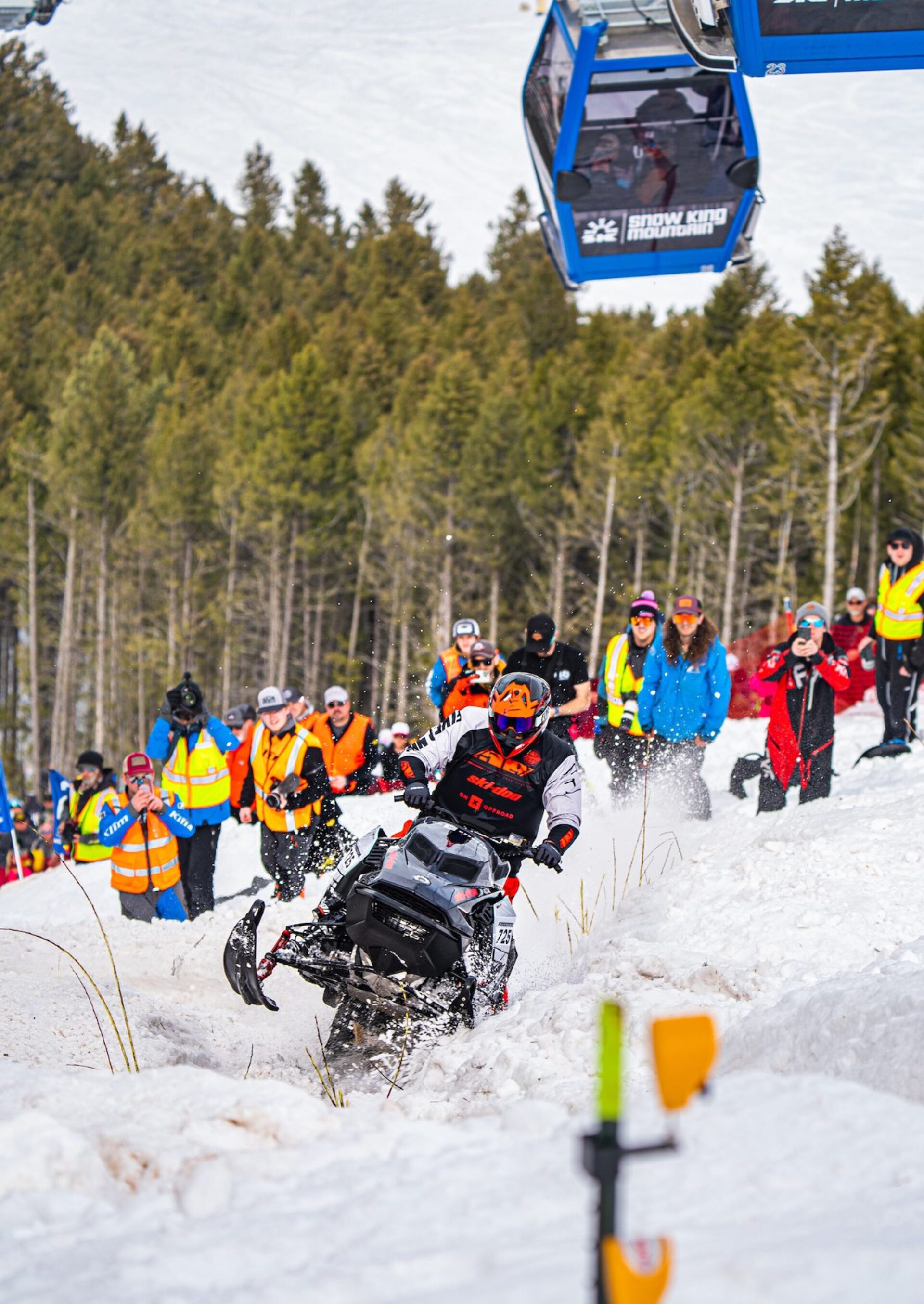 A snowmobiler on the mountain, with spectators watching in the background.