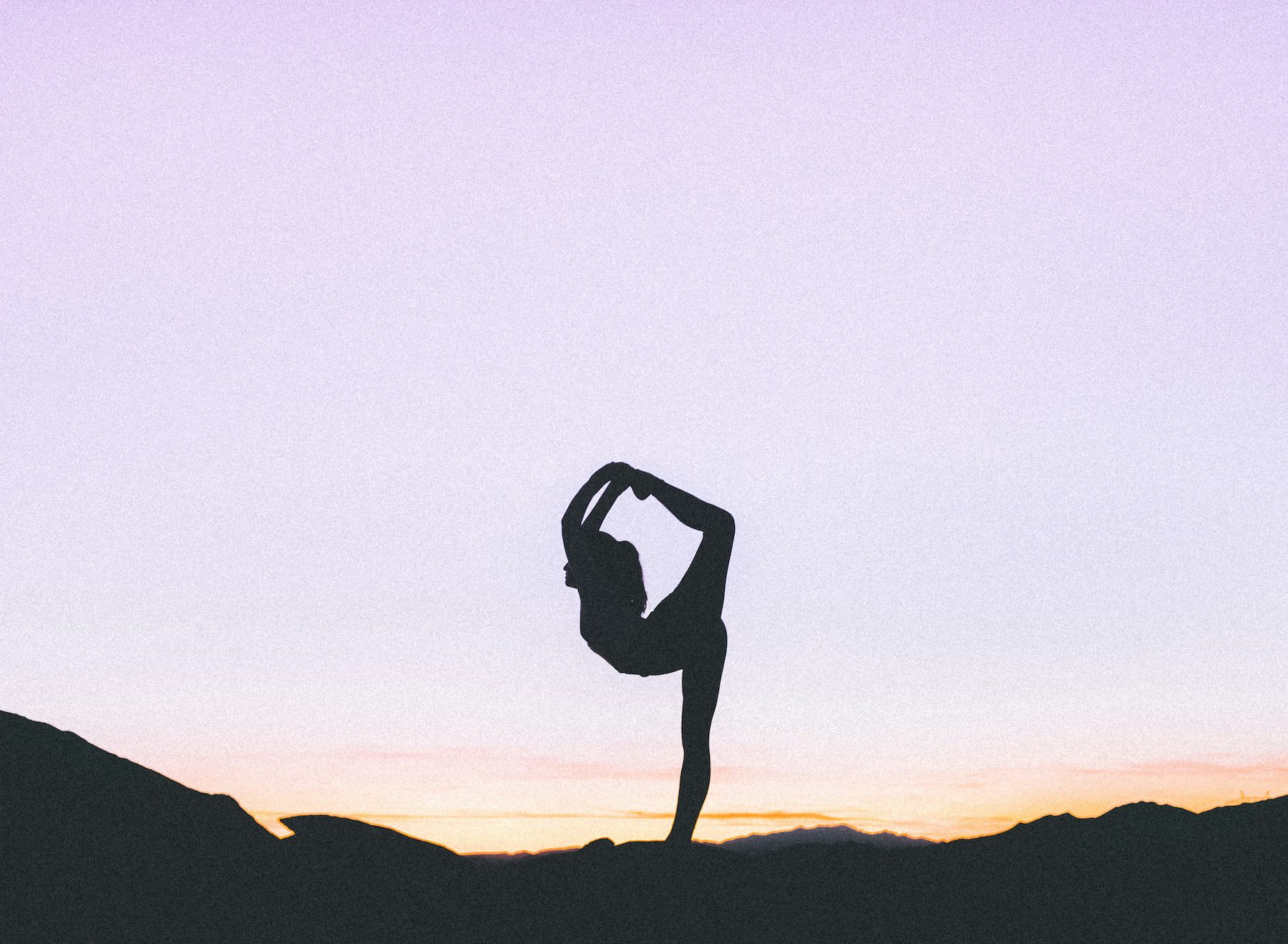A woman practicing yoga. Standing with her one leg stretched over her back. On a mountainside.