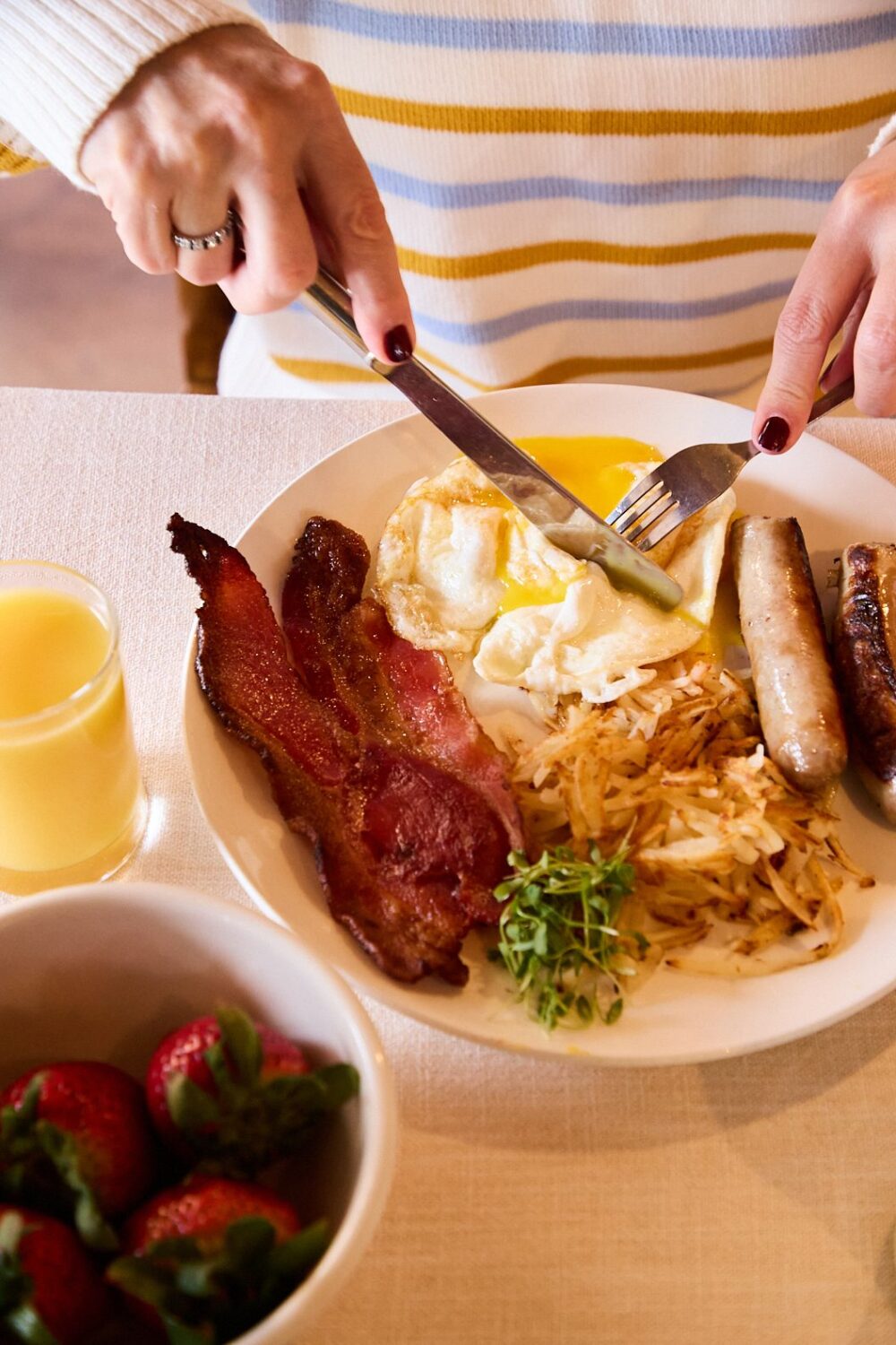 A homemade breakfast of eggs, bacon, sausage and hash browns, all with a bowl of strawberries and a glass of OJ.